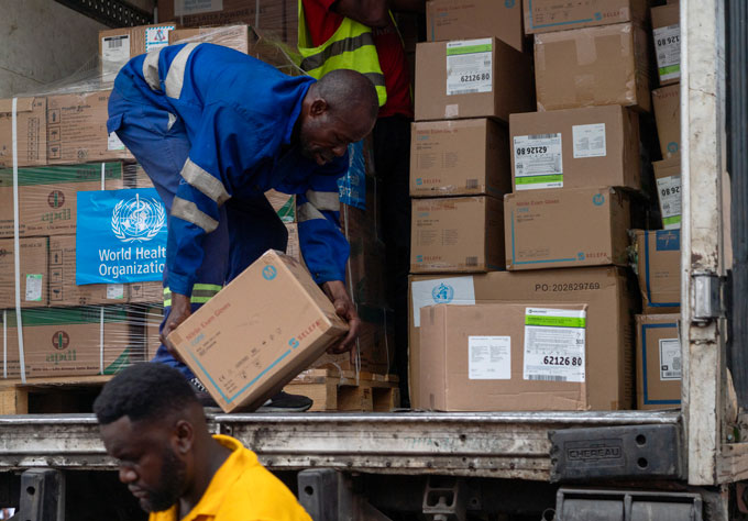 A worker unloads cardboard boxes containing medical supplies from a delivery truck in the Democratic Republic of Congo while another worker walks in the foreground.